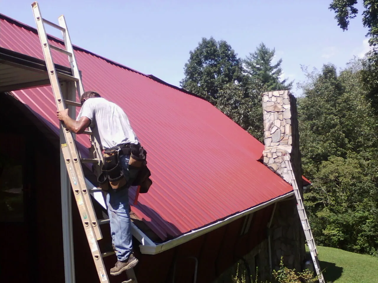 Roofer on a ladder beside a red metal roof and stone chimney — Soffit & Fascia Repair by Roofing Brothers in Elberfeld